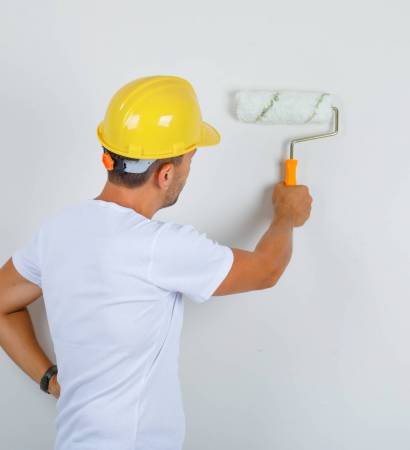 Builder man painting wall with roller in white t-shirt, helmet and looking busy , back view.